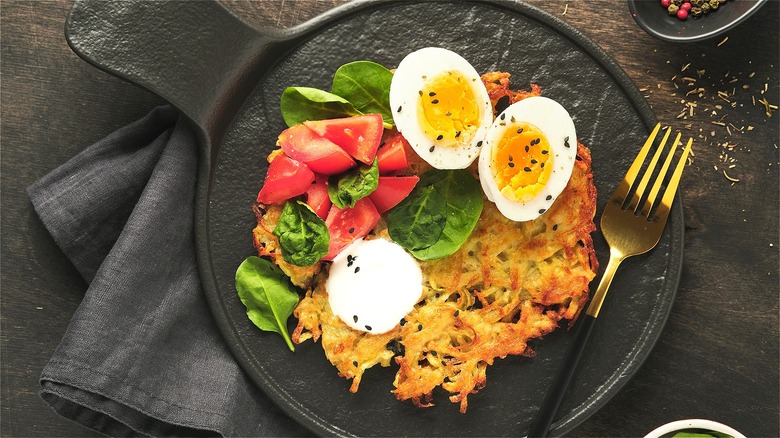 Latke served on a black plate with sour cream, chopped tomatoes, spinach, and a halved soft-boiled egg