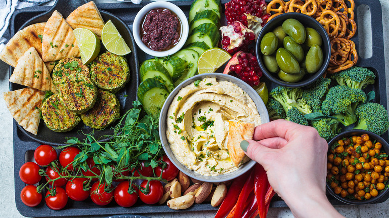 A hand dips a wedge of pita bread into a bowl of hummus on a vegan charcuterie board.