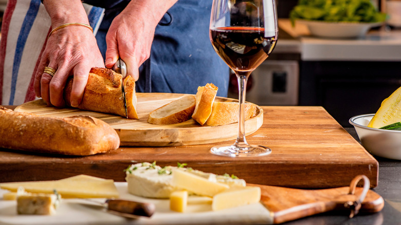 Hands are seen using a knife to slice a baguette on a wooden board, with a glass of wine and a cutting board holding various cheeses.