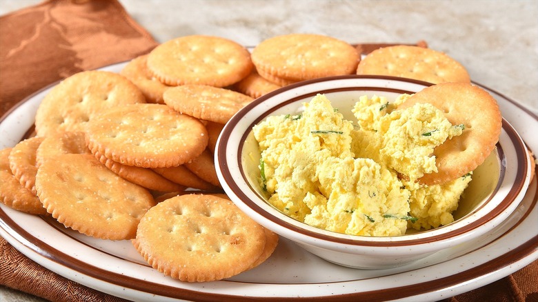 A bowl of deviled eggs dip with golden round crackers sit on a plate.