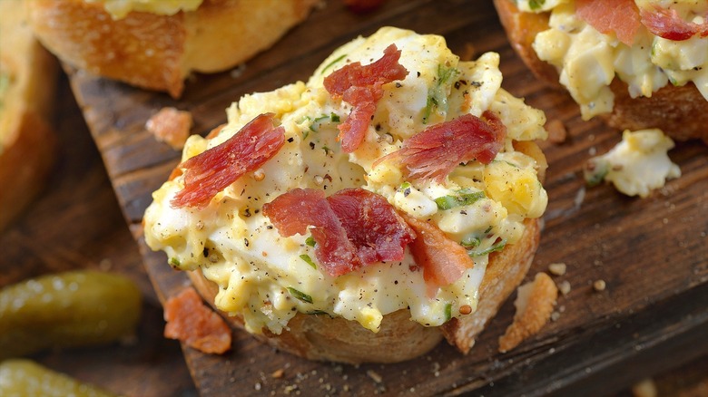 Deviled egg dip and chopped bacon are shown on a piece of crostini.