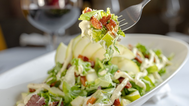 Mixed salad with bacon bits in an oval white plate with a fork holding up a mouthful of salad and bacon bits