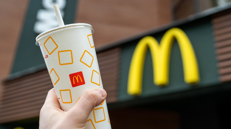 A hand holding up a McDonald's soft drink with a restaurant in the background