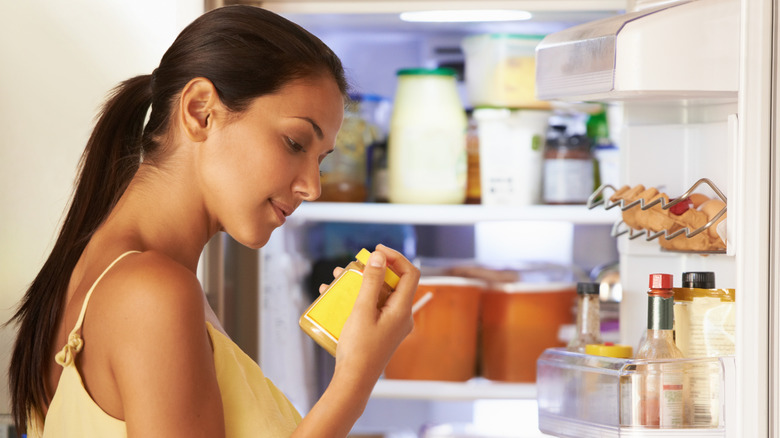 Woman looking at a jar of mustard in her open refrigerator