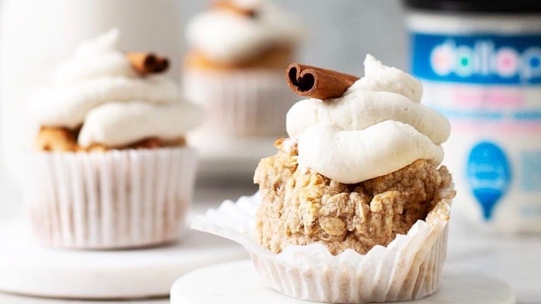 A trio of streusel cupcakes are topped with dollop icing and a cinnamon stick, with a jar of dollop in the background