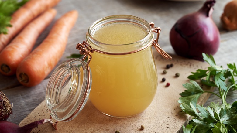 A jar of stock next to carrots, onion, and parsley