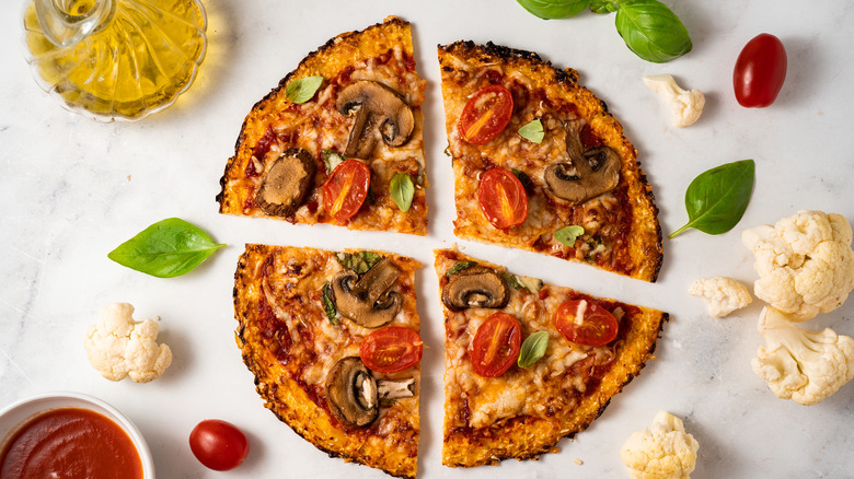 Cauliflower crust pizza cut in four pieces, surrounded by basill, cauliflower, tomatoes, jar of oil, and cup of tomato sauce