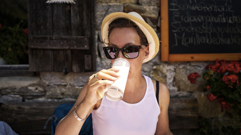 A woman in a hat with sunglasses drinks a cool glass of milk in front of a cafe