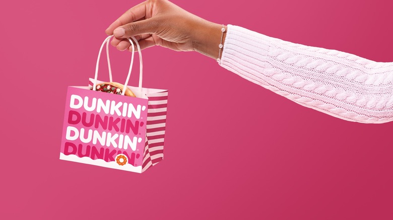 Woman's hand holding a holiday Dunkin' bag with a single donut inside on a pink background