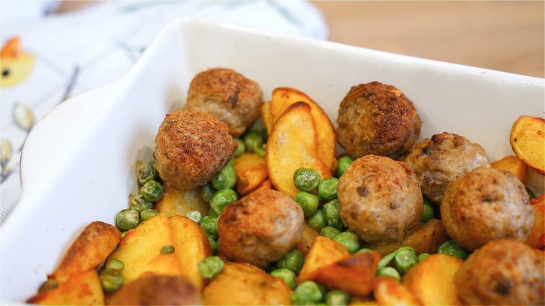 Cooked meatballs, potato wedges, and green peas in a white baking pan