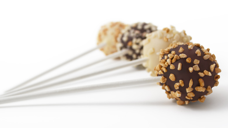 Four cake pops with nuts are lined up on a white surface.