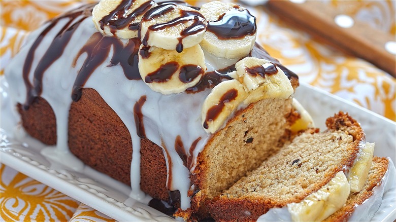 Semi-sliced loaf of banana bread covered in white icing, chocolate sauce, and fresh bananas