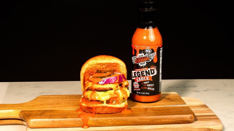 A burger on a wooden board next to a bottle of Everything Legendary's sauce