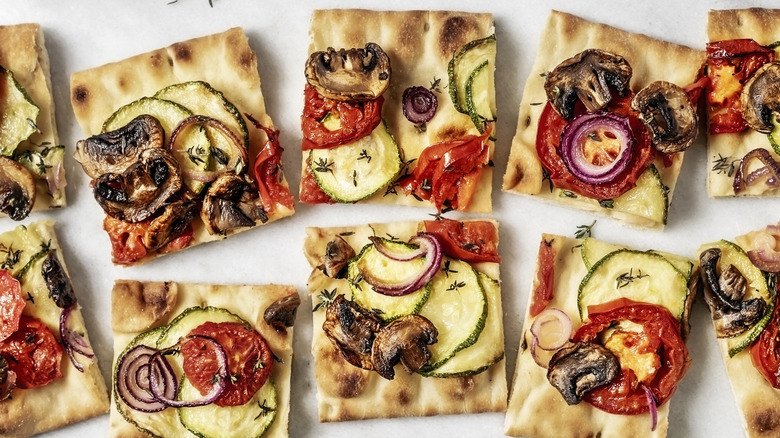 Square slices of flatbread with baked zucchini, mushroom, tomato, and onion sit on a white table