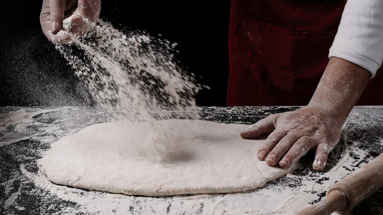 A pair of chef's hands tosses flour onto a round of pizza dough