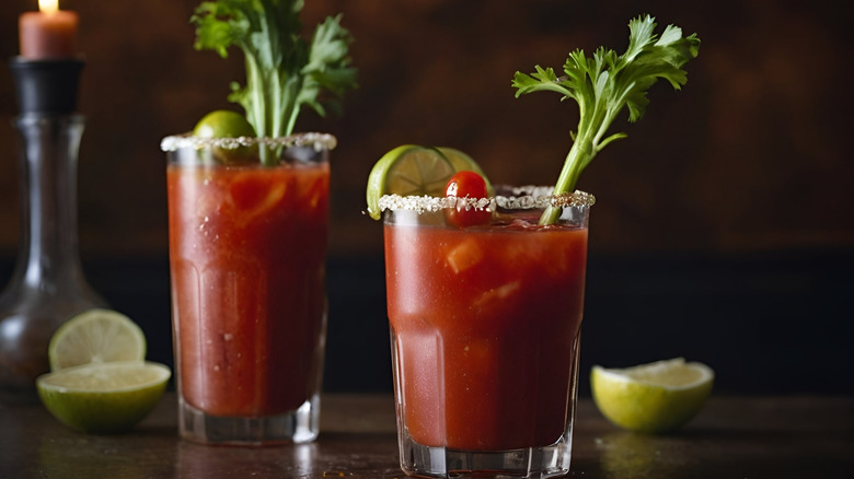 Two bloody marys with celery, cherry tomato, and lime garnishes on a wooden surfaces with cut limes and a candle in the background