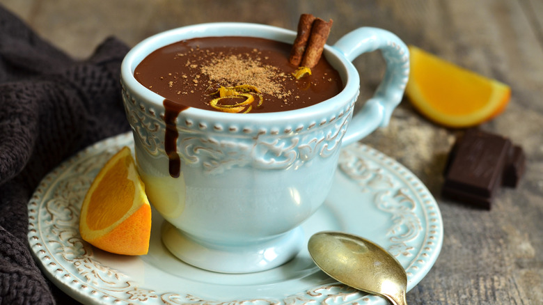 Blue-green mug topped with cinnamon, orange zest, and chocolate shavings on a wood tray with a slice of orange and spoon