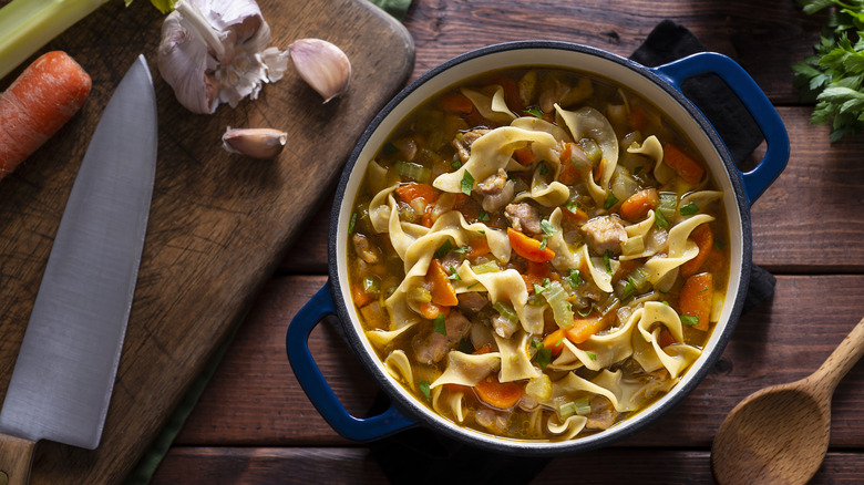 blue pot of chicken noodle soup next to knife and wooden spoon