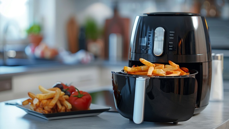 Black air fryer with basket full of fries next to plate of fries and whole tomatoes