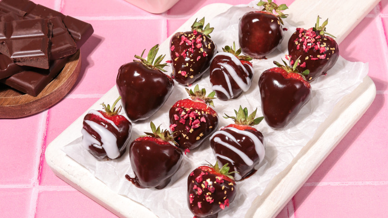 White tray of chocolate covered strawberries decorated with swirls of white chocolate and crisp decor on a pink tiled background with squares of chocolate on the side