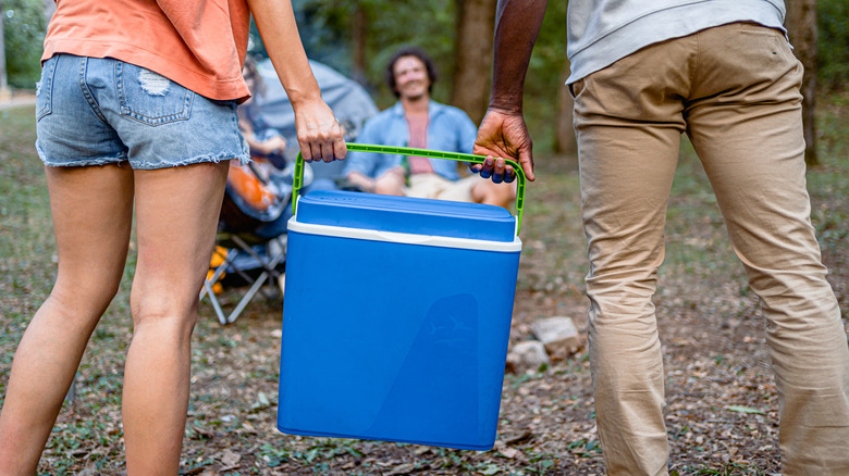 Two people carry a blue ice chest at a campsite, each with one hand on the handle.