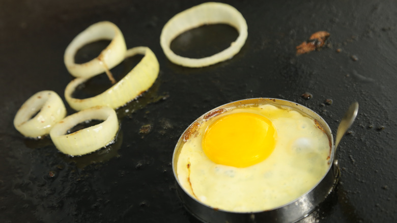 A fried egg cooking next to onions on a griddle