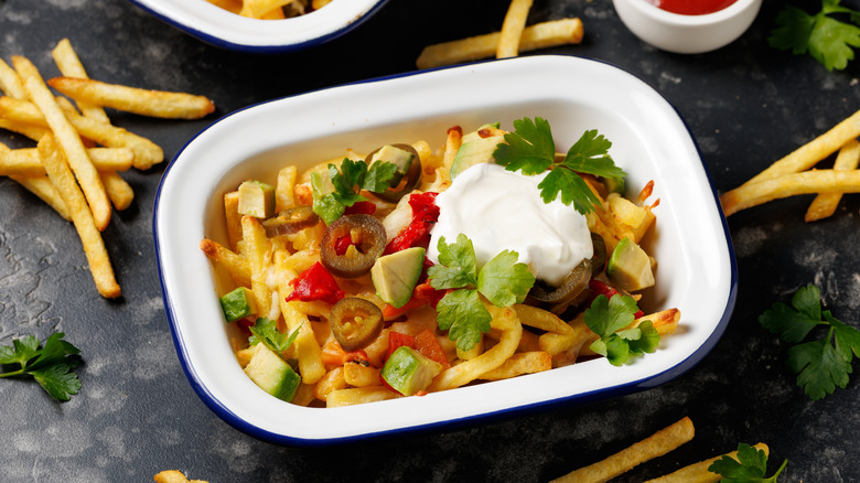 A plate of loaded french fries consists of golden fries, salsa, jalapenos, avocado, sour cream, cheese, and a fresh herb garnish