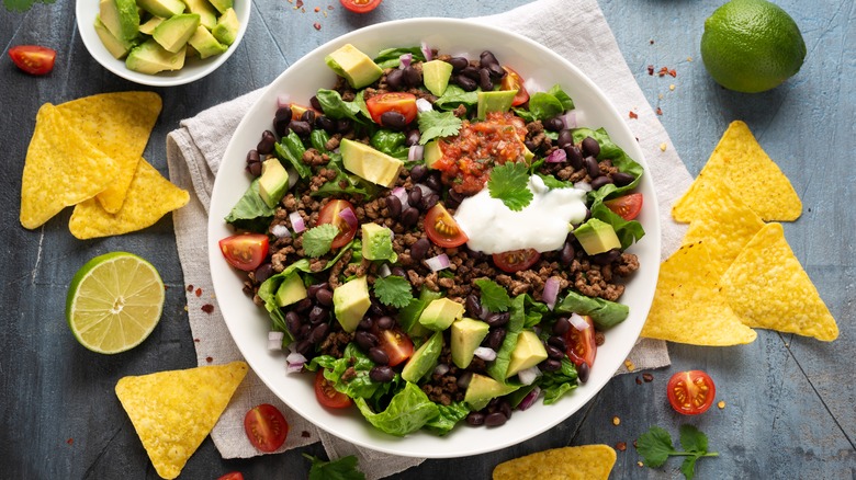 A white bowl filled with Tex-Mex salad, with tortilla chips, limes, cherry tomatoes, and a bowl of diced avocado on the counter adjacent