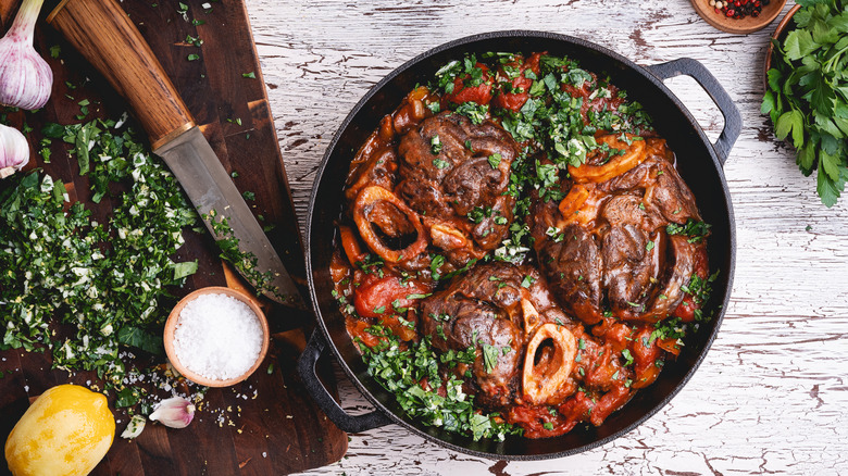 Braised meat in a Dutch oven next to a cutting board with garlic, parsley, salt, and lemon