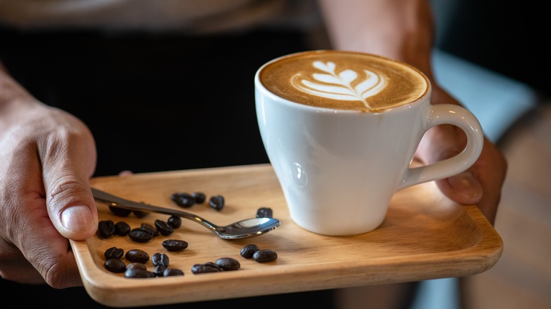 Coffe with floral milk design on top in a white cup presented on a wooden tray with small spoon