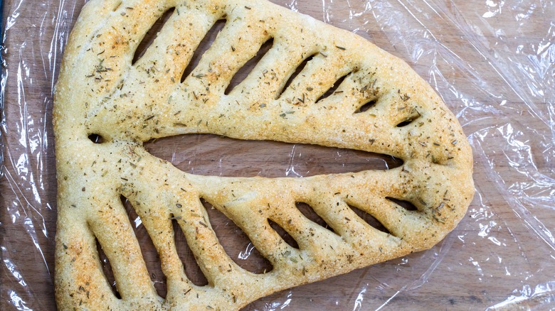 Fougasse bread topped with herbs resting on plastic wrap