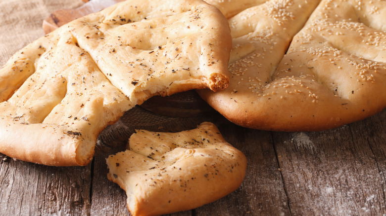 Two pieces of fougasse topped with herbs and sesame seeds sitting on a wooden board