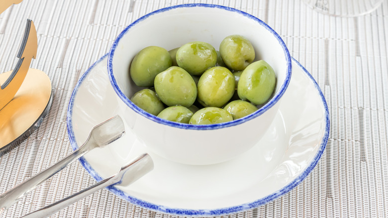 Large green Castelvetrano olives in a white bowl with serving tongs