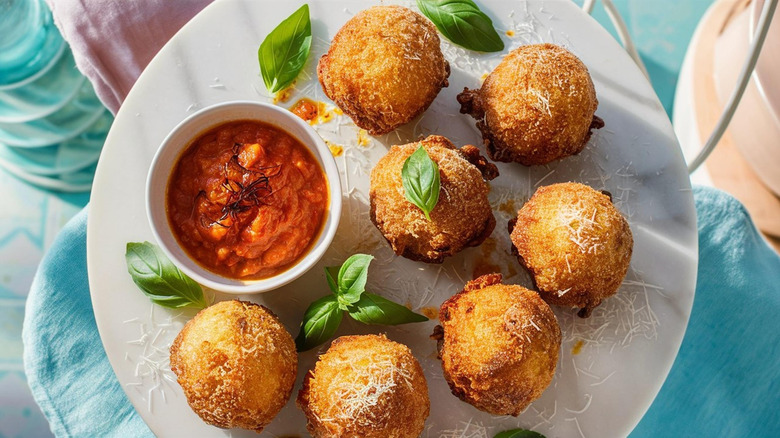 Several golden round fried croquettes on a white plate with red dipping sauce in a small bowl and scattered green basil leaves