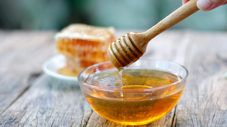 A honey wand dripping honey into a clear bowl with honeycomb in the background