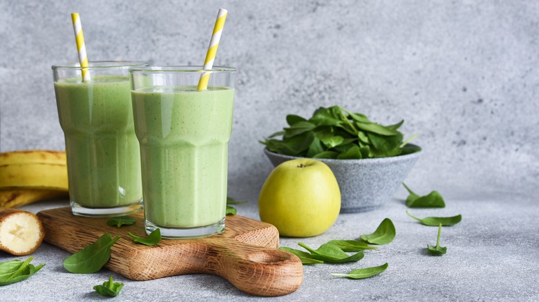 Two green smoothies on wooden board next to spinach and green apple