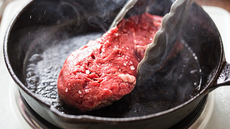 Raw steak being placed in a hot pan with tongs