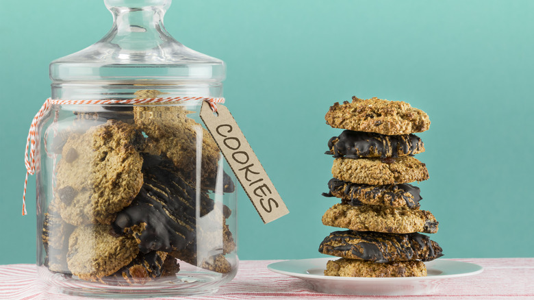 A glass cookie jar filled with cookies next to a stack of cookies in a bowl