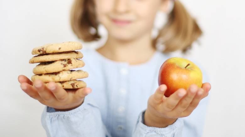 Child with pigtails holds a stack of chocolate chip cookies in one hand and an apple in the other