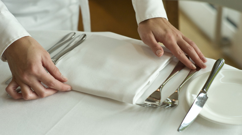 A waiter places a napkin at a table next to a fine dining place setting at a restaurant