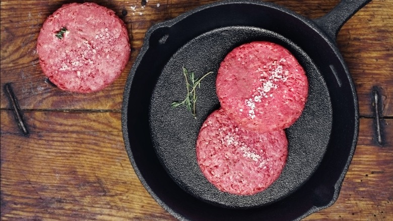 Two burger patties in a cast iron pan on a rustic wooden table next to a third patty