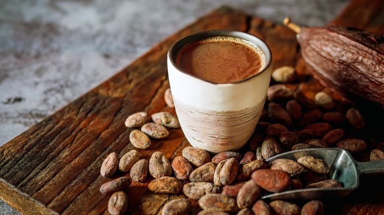 Hot cocoa drink in small ceramic cup on a wooden table and texturized table. Natural cocoa beans surrounding cup, with small metal scoop.