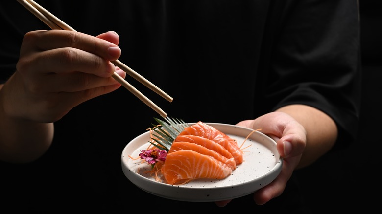 A man's hand holds a pair of chopsticks in one hand and a plate of raw salmon in the other