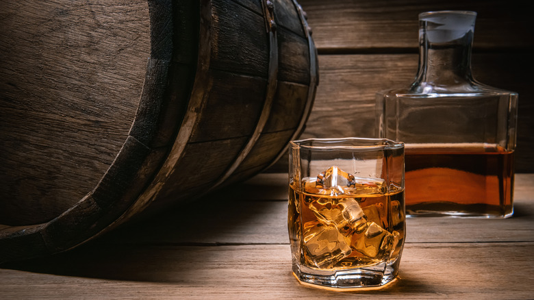 wooden whiskey barrel to the left of a whiskey-filled rock glass and bottle of whiskey