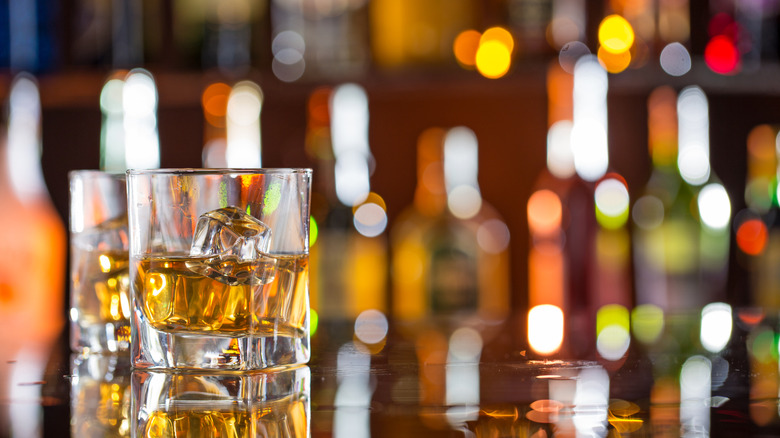 Two glasses of bourbon with ice on a bar with a variety of bottles blurred in the background