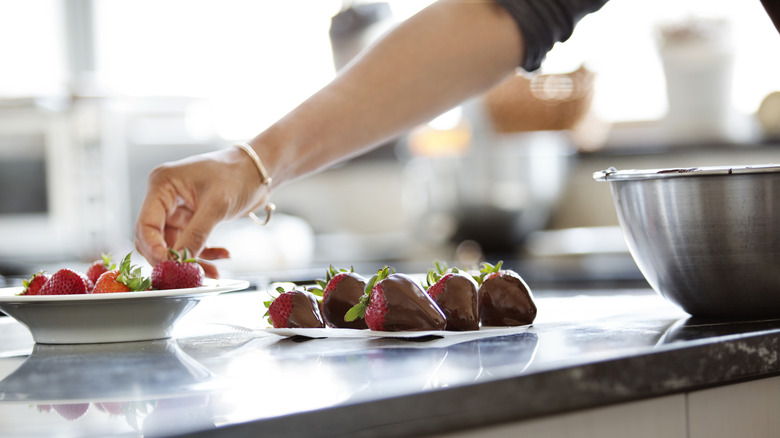 A tray of chocolate covered strawberries dries on a counter as a baker dips a fresh strawberry into melted chocolate