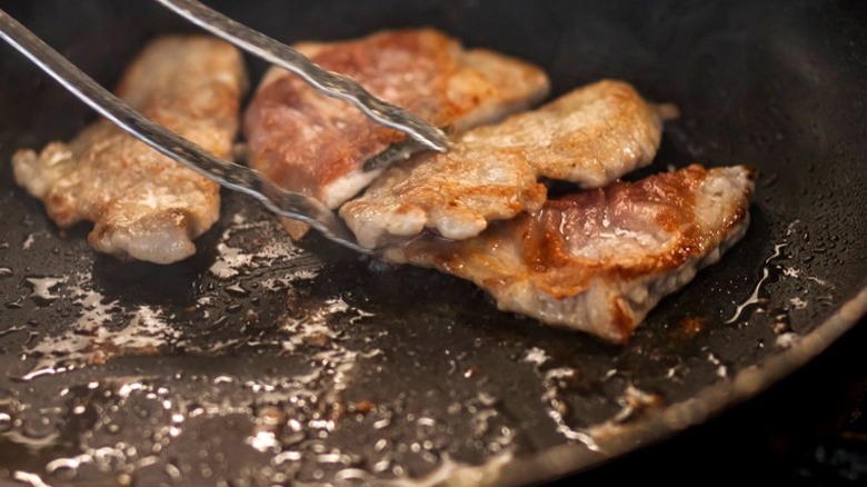 Pieces of meat being tossed with tongs while browning in pan full of oil