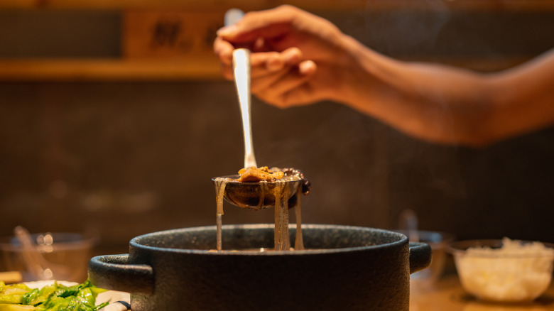 Hand holding a ladle over a pot of stew sitting on a table full of ingredients