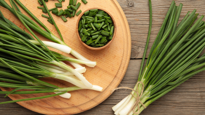 Bunches of green onions are on a wooden cutting board along with a wooden ramekin containing chopped green onion