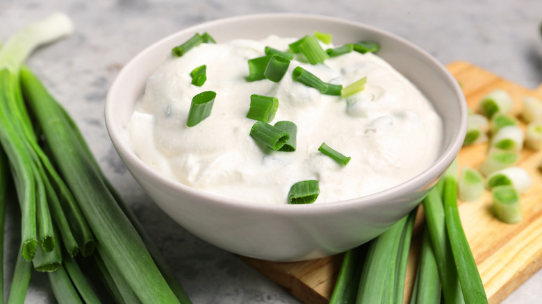 A white bowl of sour cream topped with chopped green onion sits on a wooden cutting board, with stalks of full green onions sitting on either side